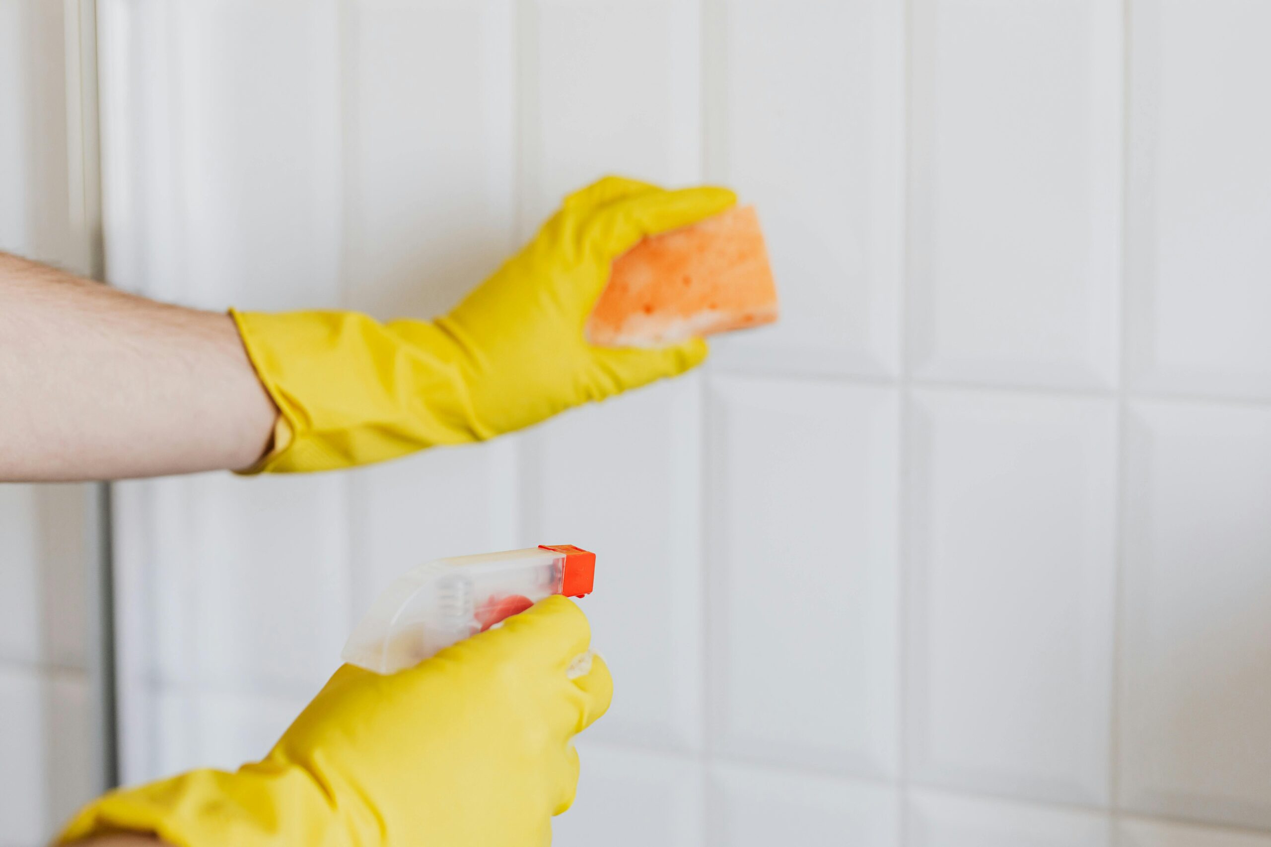 commercial-plumbing A person wearing yellow gloves cleans bathroom tiles with a sponge and spray bottle.